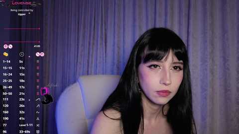 Video of an East Asian woman with long black hair and pale skin, sitting in a white chair, wearing a red dress, against a grey curtain background.