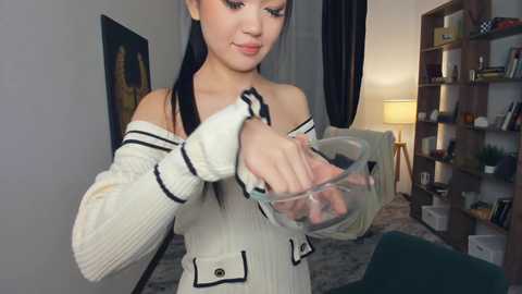 Video of an East Asian woman with long black hair, wearing a white off-shoulder sweater, holding a glass bowl, in a cozy living room with a bookshelf, a lamp, and a green chair.