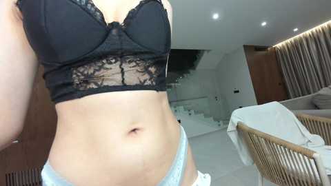 Video of a woman wearing black lace bra and light blue panties, standing in a modern, minimalist bedroom with white walls, wooden accents, and a bed.