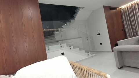 Video of a modern, minimalist bedroom with a wooden wall, glass staircase, and a gray armchair. The room features white walls, a light gray carpet, and a beige wooden headboard.