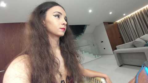 Video of a young woman with long, wavy brown hair, wearing red lipstick, sitting in a modern, minimalist living room with a grey couch and white walls.