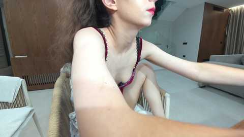 Video of a light-skinned woman with long dark hair, wearing red lace lingerie, sitting on a wicker chair, capturing a selfie in a modern, minimalist living room.