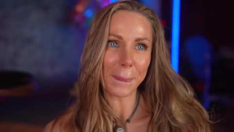 Video of a smiling, light-skinned, blonde woman with blue eyes, wearing a necklace, in a dimly lit, colorful room with blurred background.