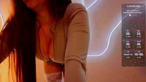Video of a partially visible woman with long dark hair, wearing a low-cut beige top, standing indoors with a glowing neon sign in the background.