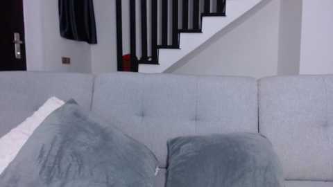 Media: Video of a modern living room with a light grey sectional sofa, featuring soft, plush cushions in a similar shade. A staircase with black railing and white walls is visible in the background.