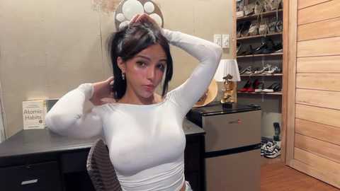 Video of a woman with medium-sized breasts in a tight white top, adjusting her hair in a dressing room with a mirror, wooden shelves, and a shoe rack.