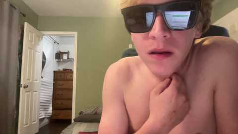 Media: Video of a topless young woman with fair skin, wearing large black 3D glasses, standing indoors. She has short, blonde hair and a shocked expression. The room has beige walls, a white door, and a wooden dresser.