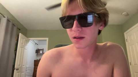 Media: Video of a young, fair-skinned, shirtless man with short, light brown hair, wearing large, dark sunglasses indoors. Background features a closed door, a gray curtain, and a wooden cabinet.