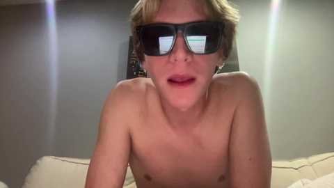 Media: Video of a shirtless, pale-skinned, young man with short blonde hair, wearing black 3D glasses, lying on a beige couch in a dimly lit room.
