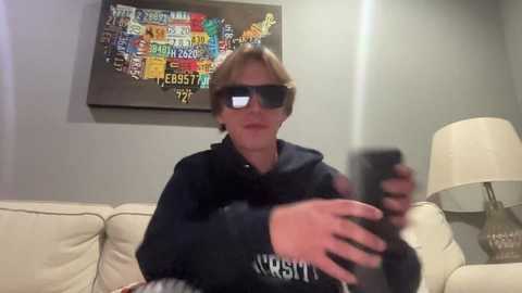 Media: Video of a blonde woman in dark glasses and a black hoodie, holding a phone, seated on a beige sofa. A colorful, abstract wall art with text hangs behind.