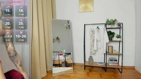 Video of a modern, minimalist bedroom with a mirror, a black-framed clothing rack, beige curtains, and wooden floors. A woman with fair skin and long hair in a red top is partially visible.