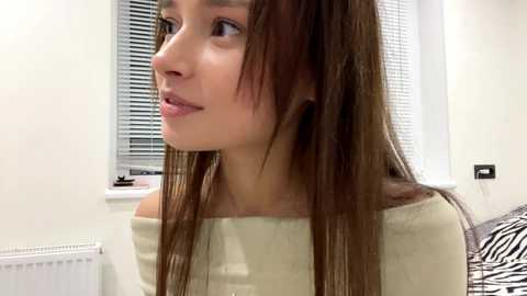 Video of a young woman with long brown hair, fair skin, and a light beige sweater, looking to the side in a modern, white-walled bedroom with closed blinds and a zebra-patterned pillow.