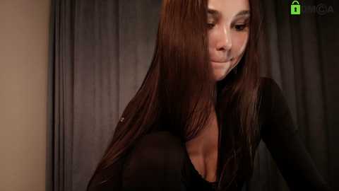 Video of a woman with long, straight brown hair, wearing a black top, standing in front of dark curtains. Her face is partially obscured by her hair.