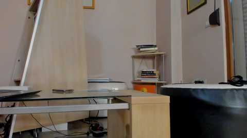 A video of a cluttered, small room with a glass desk, wooden shelves, and various tools and materials scattered about, including a ladder and a black bag. The walls are light pink with framed art.