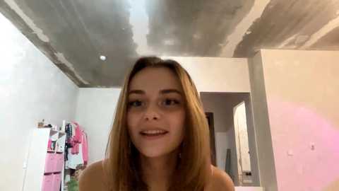 Video of a young woman with light skin and long, straight brown hair, smiling, in a partially renovated room with exposed concrete ceiling, pink clothes on racks in background.