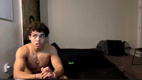 Video of a shirtless, curly-haired young man with medium skin tone lying on a bed in a sparsely furnished, dimly lit bedroom.