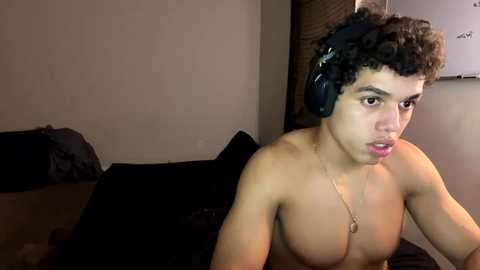 Video of a shirtless, young Hispanic man with curly black hair and medium skin tone, wearing large black headphones, sitting on a black bed in a dimly lit bedroom.