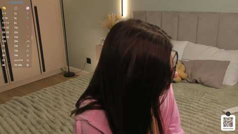 Media: Video of a young girl with long, dark hair, wearing a pink hoodie, facing away from a modern, beige-tufted bed with a plush teddy bear and pillows. The room has light wooden floors and a sliding door.