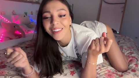 Video of a young Latina woman with long dark hair, wearing a white t-shirt, lying on a floral bedspread. She smiles, holding a phone. Pink fairy lights in the background.