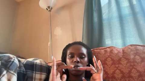 Video of a woman with dark skin, long black hair, and light makeup, lying on a patterned couch, applying lipstick. Background includes a beige wall, a lamp, and a teal curtain.