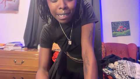 A video of a young Black woman with dreadlocks and a black outfit, holding a black garment. The background features a wooden dresser, a red armchair, and a colorful painting.