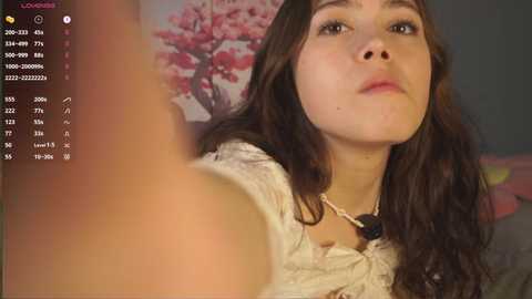 A video of a young woman with long, wavy brown hair, wearing a lace top, taking a selfie with a phone, blurred background showing a pink flower wall.