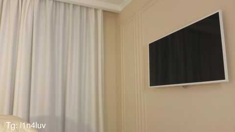 Video of a beige wall with a mounted flat-screen TV and a sheer white curtain partially covering a window. The room has a minimalist, modern aesthetic.