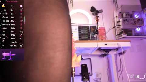 A video of a dark-skinned man in a dimly lit room with a desk featuring a computer monitor displaying a black screen with white text and a pink drink. The room is cluttered with various objects, including a microphone.