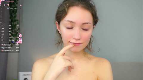 A topless young woman with fair skin, brown hair, and delicate features is seen in a live stream, finger to her lips, in a simple room with a gray wall.