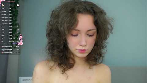 Video of a young woman with curly brown hair, light skin, and closed eyes, seated topless in a gray room. Background shows social media icons and a green plant.