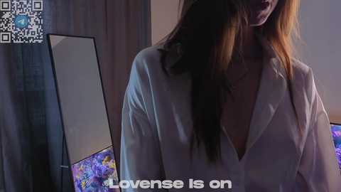 Video of a woman with long brown hair, wearing a white robe, standing in front of a mirror. The background shows a dark curtain and a colorful flower arrangement. Text reads, \"Lavense Is On.\