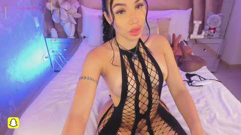 A video of a Latina woman with long black hair, wearing a black fishnet bodysuit, sitting on a bed in a dimly lit bedroom.
