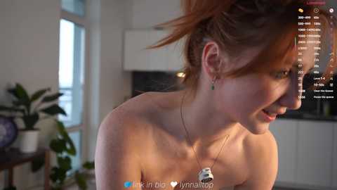 Video of a topless woman with fair skin and red hair tied up, wearing a necklace. Background shows a modern kitchen with plants and digital screens displaying social media content.