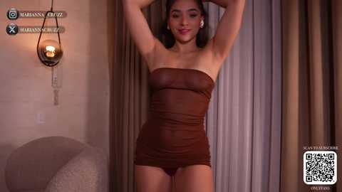 Video of a young woman with medium skin tone, wearing a sheer brown strapless mini dress, standing in a dimly lit room with beige curtains and a pendant light.