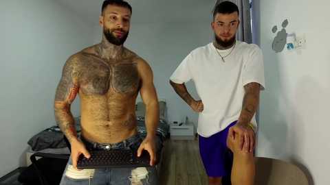 Video of two shirtless men with tattoos, one holding a keyboard, the other in shorts, standing in a minimalist room.