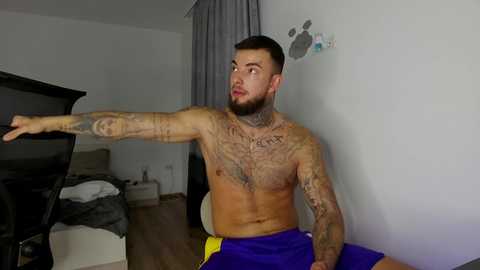 Video of a shirtless, tattooed man with short hair and beard, wearing blue shorts, pointing to a TV in a minimalist bedroom with white walls and a gray curtain.