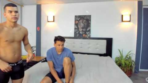 Video of a muscular, shirtless man standing on the left, a young man in a blue shirt and shorts seated on a bed in the middle, and a framed picture on the wall.