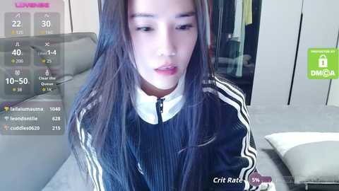 A video of a young East Asian woman with long black hair, wearing a black Adidas jacket, sitting indoors, with a digital overlay showing social media metrics.