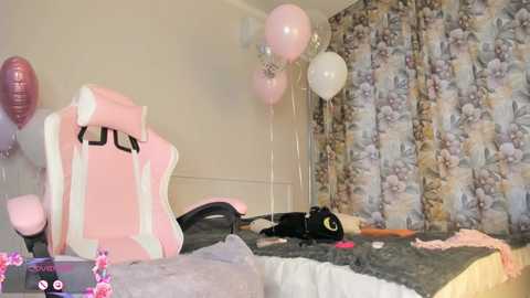 Video of a cluttered bedroom with a pink gaming chair, balloons, a black laptop, and a messy bed with floral curtains in the background.
