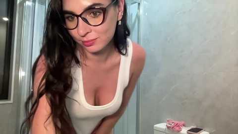 Video of a slender woman with long, dark hair, wearing black-framed glasses and a white tank top, leaning forward in a modern bathroom with light-colored tiles and a pink cloth.