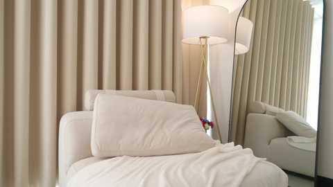 Video of a cozy, modern bedroom with beige curtains, a white chair, a white pillow, a white lamp with a white shade, and a full-length mirror reflecting part of the room.