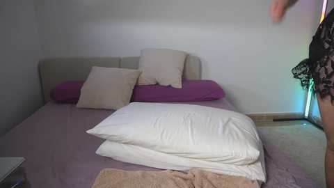 Video of a minimalist bedroom with a beige headboard, purple bedspread, and white pillows. A person in black lace lingerie stands in the corner, partially visible.