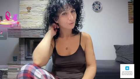 Video of a woman with curly black hair, wearing a dark tank top, sitting on a couch, looking at a phone. Background features a stone fireplace, neon lights, and a plaid shirt.