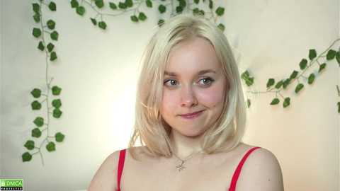 Video of a fair-skinned, blonde-haired woman with shoulder-length hair, wearing a red spaghetti-strap top, smiling, against a backdrop of green ivy vines.