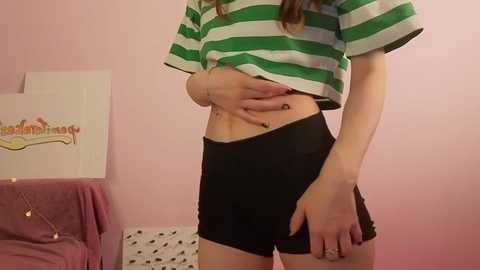 Video of a young woman with light skin and brown hair, wearing a green and white striped shirt, black shorts, and a bracelet, standing in a pink-walled room.