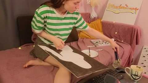 Media: Video of a young girl with light brown hair in a green and white striped shirt, sitting on a pink couch, painting a white outline of a horse on a wooden board, with a laptop, coffee mug, and paintbrushes nearby.