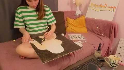 Media: Video of a young woman with long brown hair, wearing a green and white striped t-shirt, painting a guitar on a pink couch.
