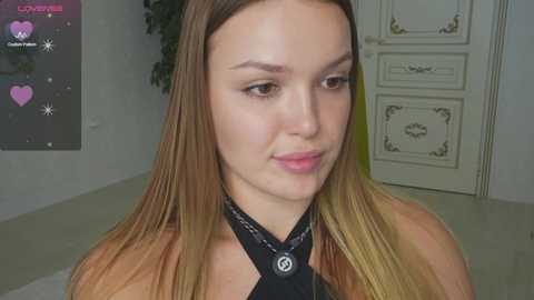 Video of a young woman with fair skin and long, straight, light brown hair, wearing a black halter top with a silver pendant. She stands indoors with a cream-colored wall and ornate white door in the background.