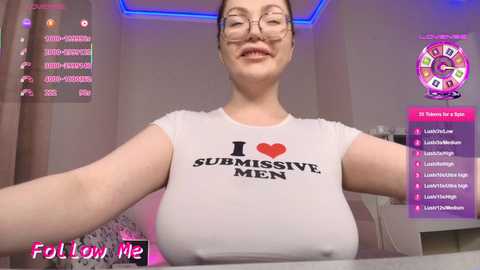 Video of a smiling, fair-skinned woman with glasses, wearing a white t-shirt with \"I \u2665 SUBMISSIVE MEN\" and a red heart. Background includes a pink room with a blue LED strip, a chat window, and a pink banner with icons.