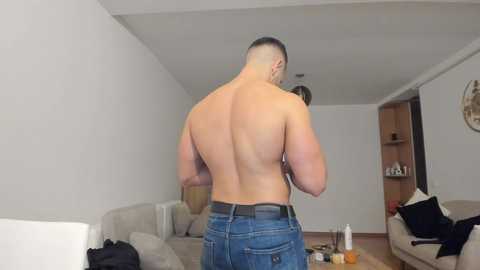 Video of a muscular, shirtless man with short hair, wearing blue jeans, standing in a modern, minimalist living room with light-colored furniture and neutral decor.